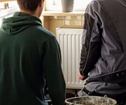 Zwei Handwerker knien in einer Küche vor einer Heizung und bereiten ihre Arbeit vor