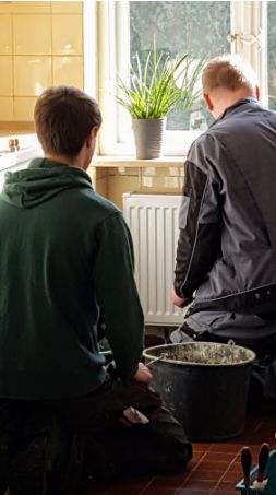 Zwei Handwerker knien in einer Küche vor einer Heizung und bereiten ihre Arbeit vor